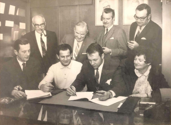 a group of people standing around a man signing a document at a desk